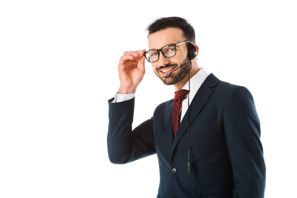 Lächelnder Callcenter-Betreiber mit Headset, der in die Kamera schaut und Brille anfasst, isoliert auf Weiß - Foto, Bild