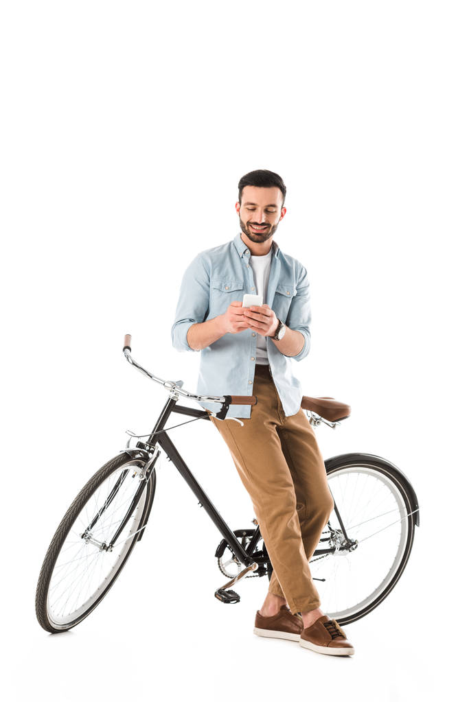 όμορφος άντρας με γένια με ποδήλατο χαμογελαστός ενώ χρησιμοποιεί smartphone απομονώνεται σε λευκό - Φωτογραφία, εικόνα