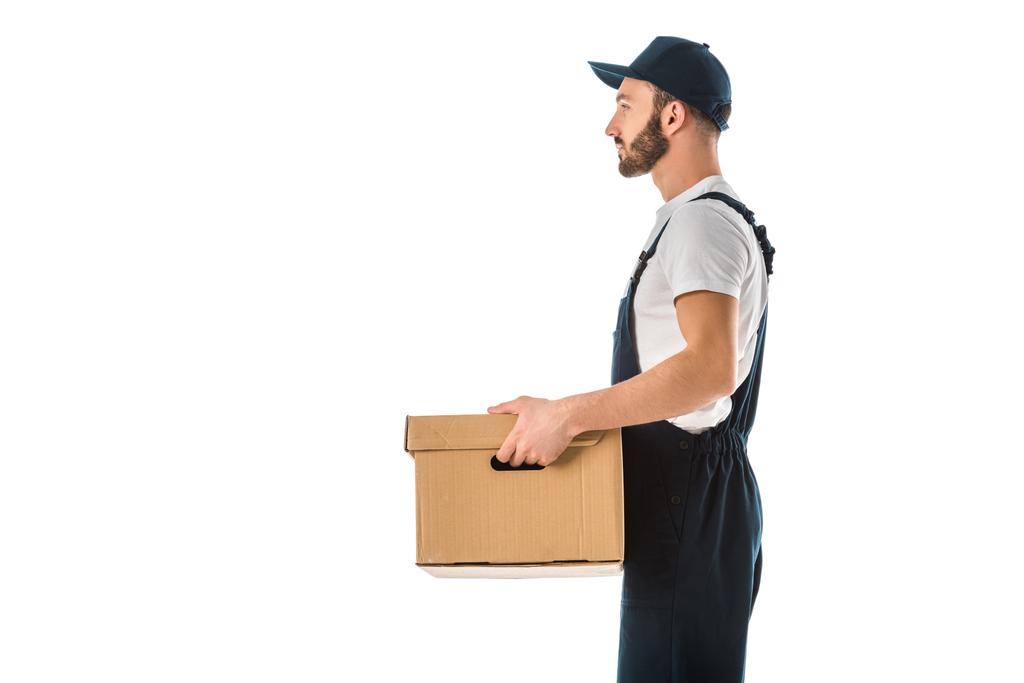 Zijaanzicht van de levering man in overalls en Cap Holding kartonnen doos geïsoleerd op wit - Foto, afbeelding