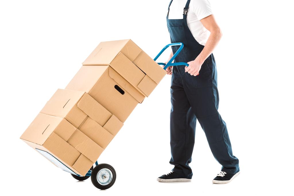 vue recadrée du livreur en salopette transportant un camion à main chargé de boîtes en carton isolées sur blanc
 - Photo, image