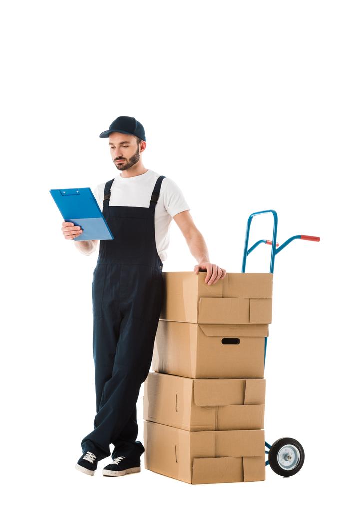 serieuze levering man staande in de buurt van handtruck geladen met kartonnen dozen en kijken naar Klembord geïsoleerd op wit - Foto, afbeelding