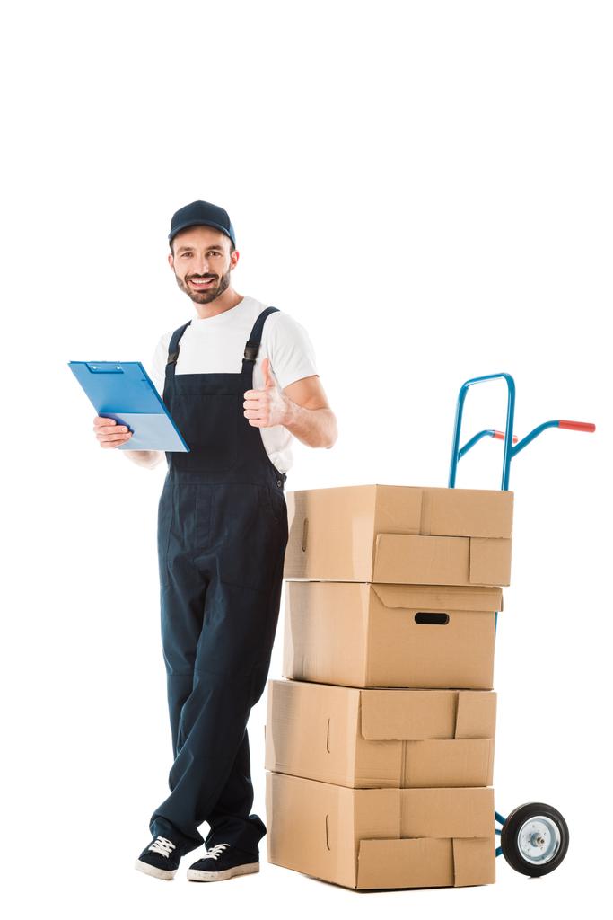 Happy delivery man staande in de buurt van handtruck met kartonnen dozen en tonen duim omhoog geïsoleerd op wit - Foto, afbeelding
