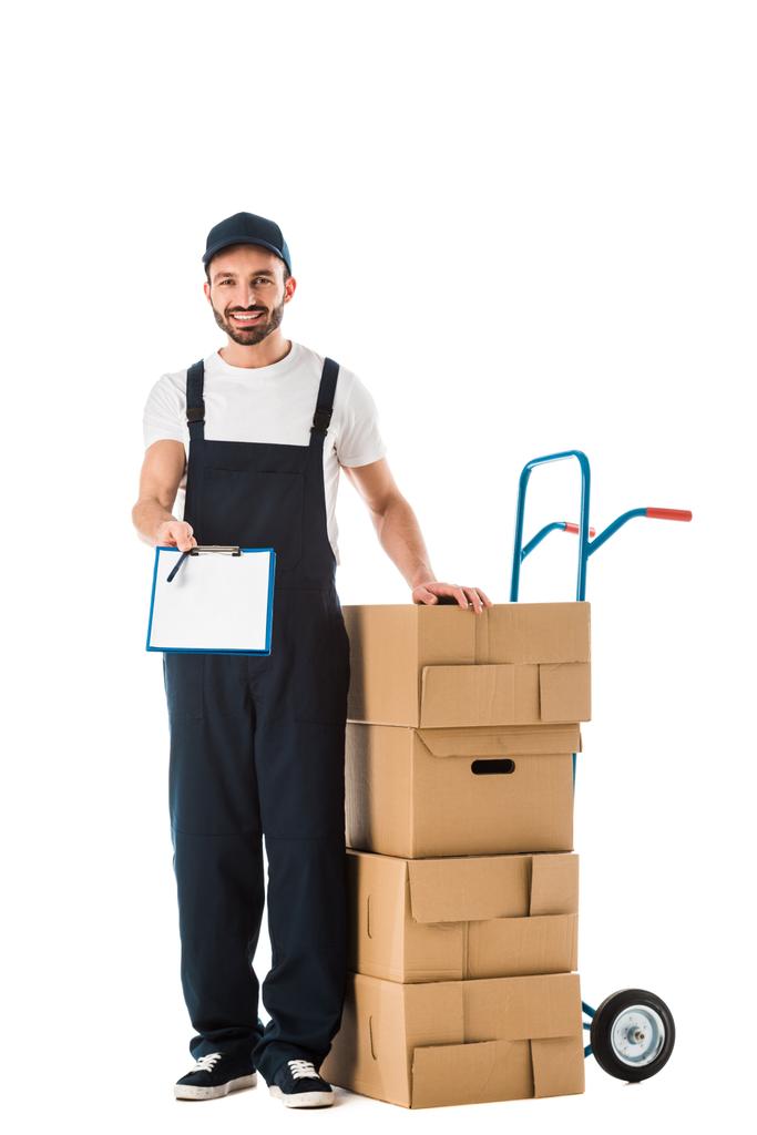 smiling delivery man holding clipboard with blank screen while standing near hand truck loaded with cardboard boxes isolated on white - Photo, Image