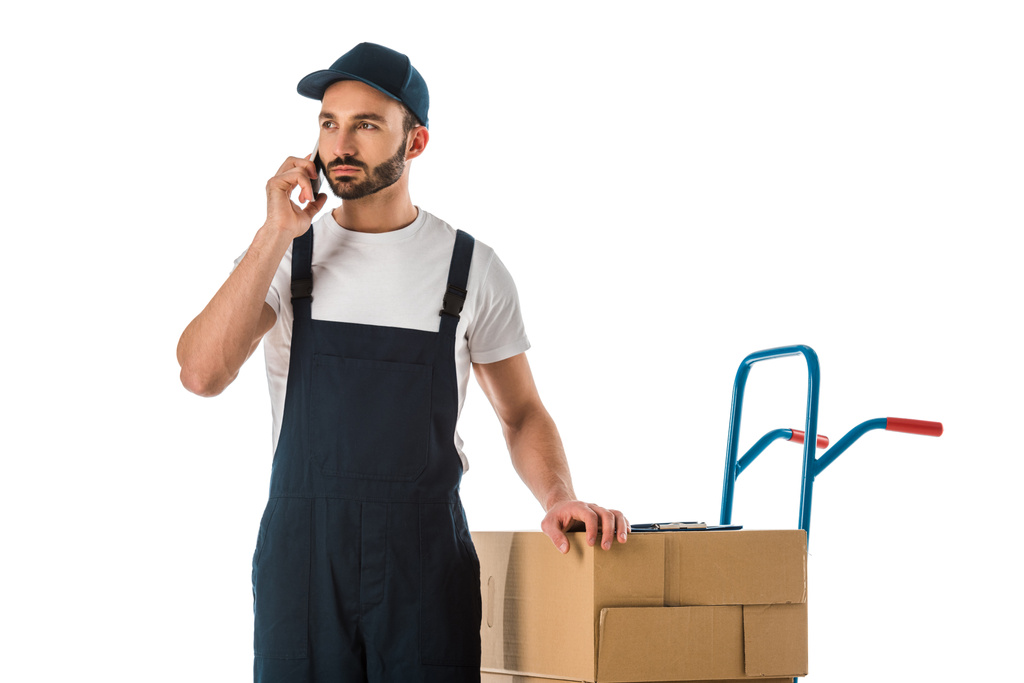 hombre de entrega seria hablando en el teléfono inteligente mientras está de pie cerca de cajas de cartón cargadas en camión de mano aislado en blanco
 - Foto, Imagen