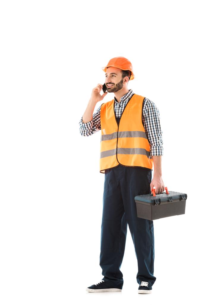 улыбающийся строитель разговаривает по смартфону, держа ящик с инструментами и отводя взгляд изолированным на белом
 - Фото, изображение