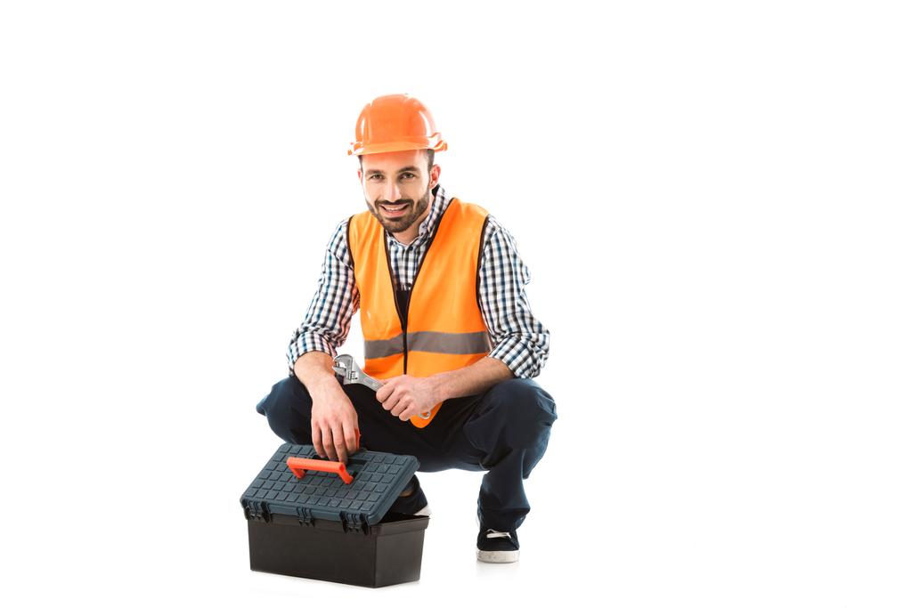 alegre trabajador de la construcción sentado cerca de la caja de herramientas y la celebración de llave ajustable aislado en blanco
 - Foto, imagen