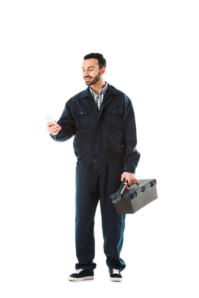 vrolijke workman in overalls met behulp van smartphone en holding Toolbox geïsoleerd op wit - Foto, afbeelding