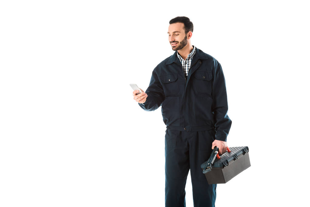 Lächelnder Arbeiter in Overalls mit Smartphone, während er Werkzeugkiste isoliert auf weiß hält - Foto, Bild