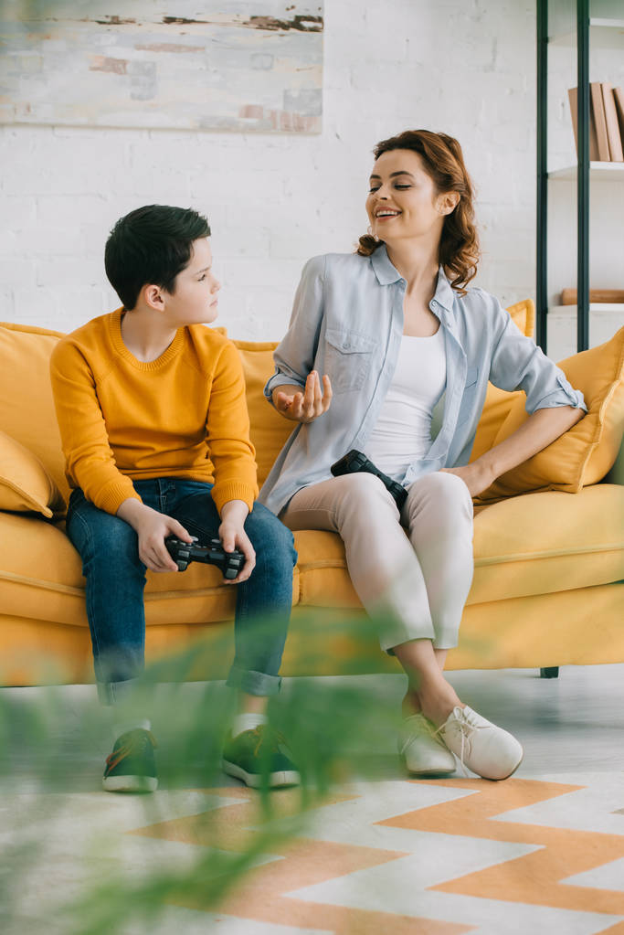 KYIV, UKRAINE - APRIL 8, 2019: Cheerful mother gesturing and looking at upset son holding joystick while sitting on sofa at home - Photo, Image