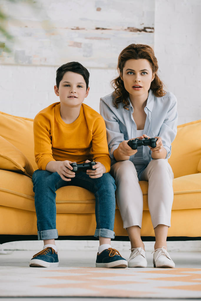 KYIV, UKRAINE - 8 de abril de 2019: Mãe e filho atentos jogando videogame com joysticks enquanto se sentam no sofá amarelo
 - Foto, Imagem