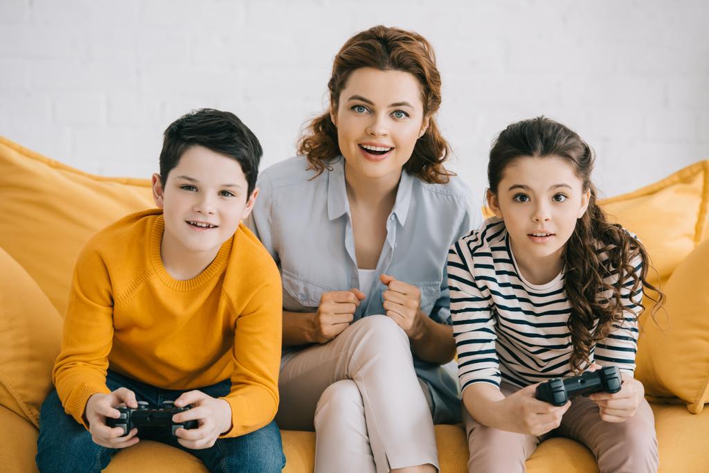 Kiev, Ukrayna - 8 Nisan 2019: Joystick'lerle video oyunu oynayan çocukların yanında kanepede oturan heyecanlı anne - Fotoğraf, Görsel