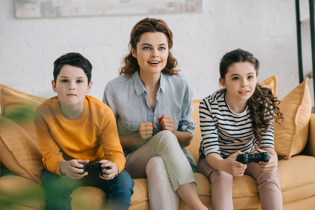 KYIV, UCRANIA - 8 de abril de 2019: Mujer alegre sentada en un sofá cerca de niños jugando videojuegos con joysticks
 - Foto, imagen