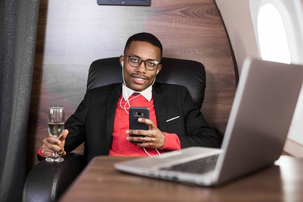 Ελκυστικός και επιτυχημένος Αφρικανός Αμερικανός επιχειρηματίας με γυαλιά που εργάζονται σε ένα φορητό υπολογιστή ενώ κάθεται στην καρέκλα του ιδιωτικού τζετ του - Φωτογραφία, εικόνα