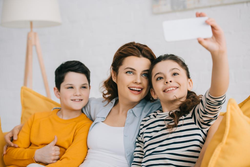 cheerful child taking selfie smiling with mother and brother while sitting on sofa at home - Photo, Image