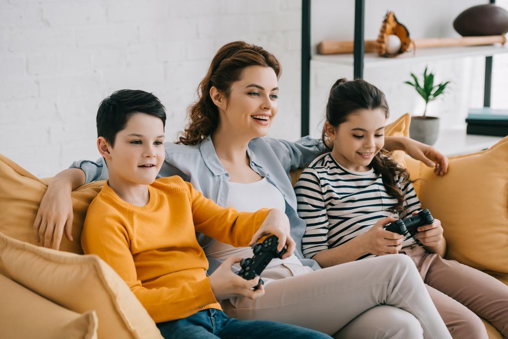KYIV, UCRAINA - 8 APRILE 2019: Donna sorridente seduta sul divano vicino ai bambini che gioca al videogioco con i joystick
 - Foto, immagini