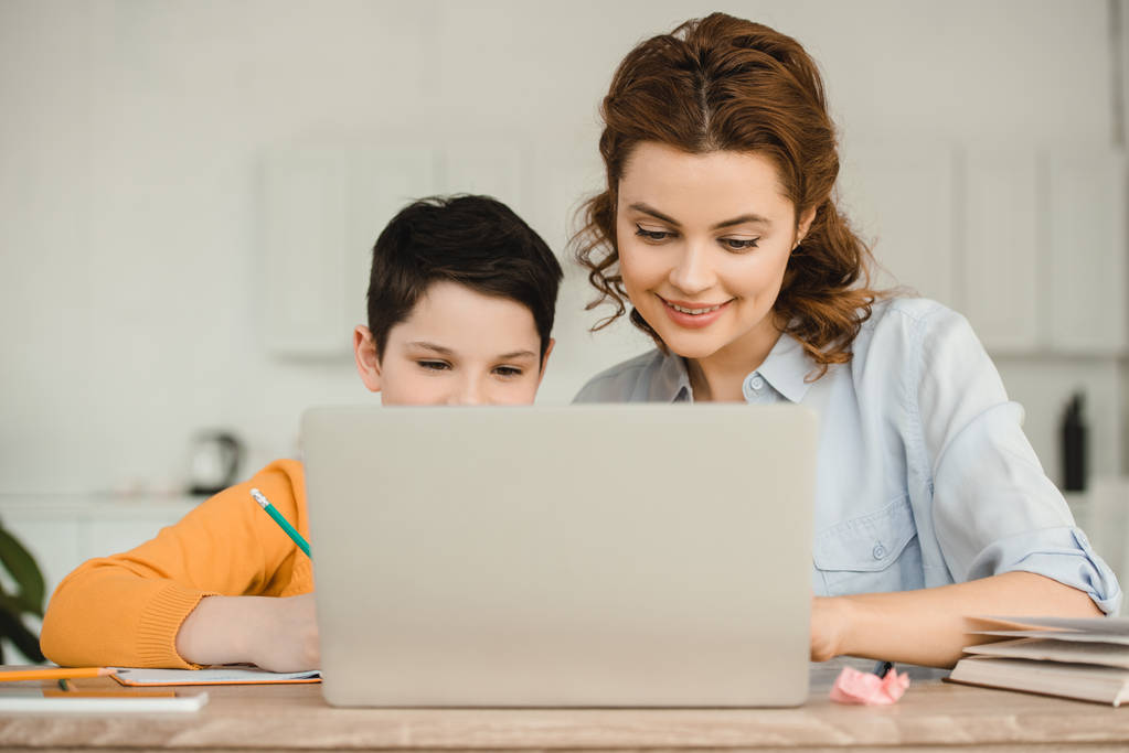 счастливая мать с милым сыном, использующим ноутбук вместе во время занятий в школе дома
 - Фото, изображение