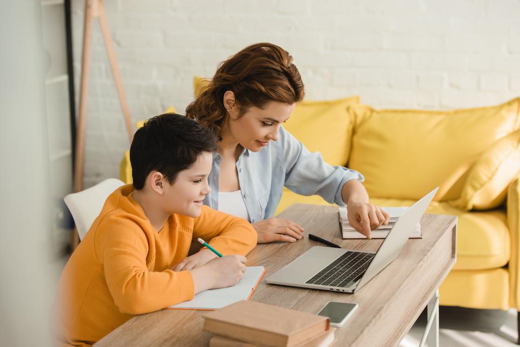 mère souriante aidant son fils attentif à faire du travail scolaire tout en étant assis au bureau avec ordinateur portable à la maison
 - Photo, image