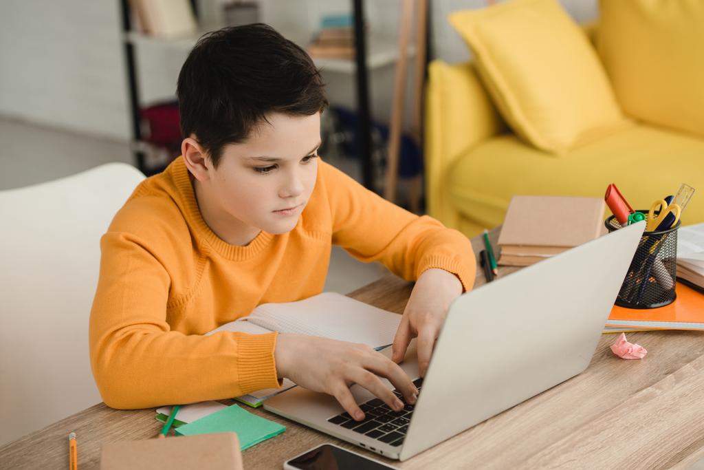 garçon attentif en utilisant un ordinateur portable tout en étant assis au bureau en bois et faire des devoirs
 - Photo, image