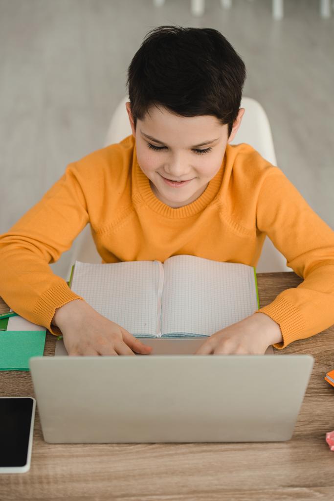 niño sonriente enfocado usando el ordenador portátil mientras está sentado en el escritorio con el libro de copia y haciendo el trabajo escolar en casa
 - Foto, imagen