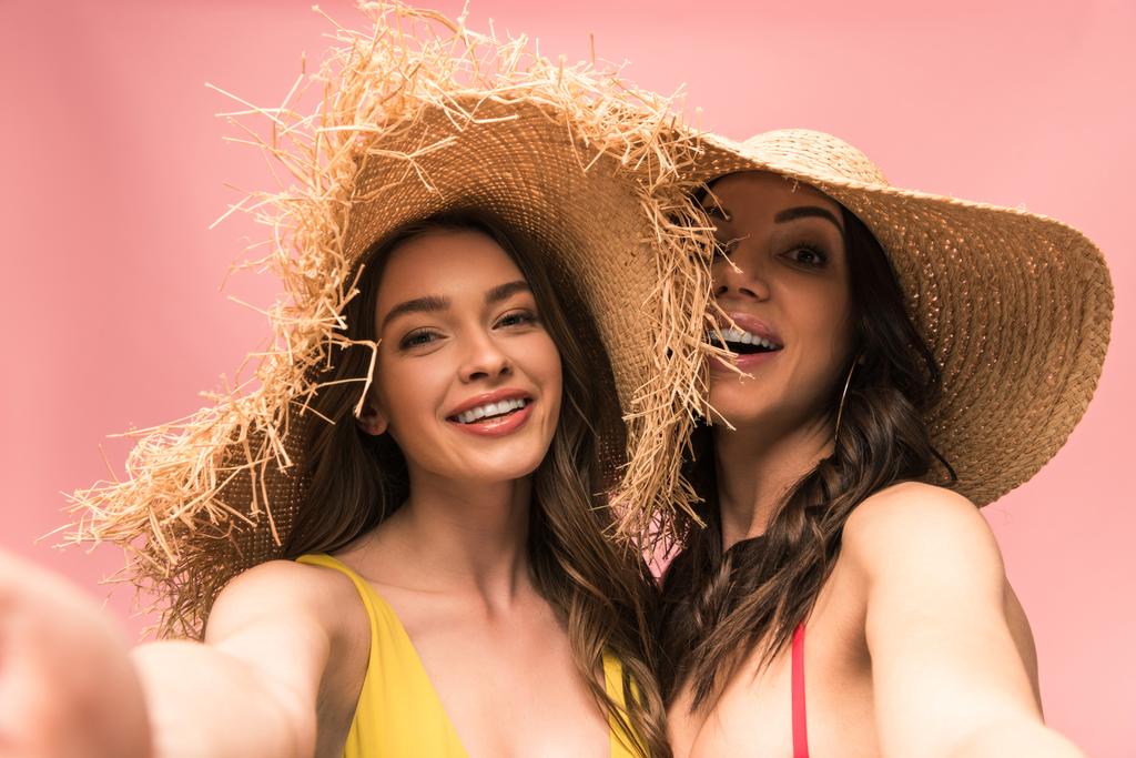 twee glimlachende meisjes in stro hoeden nemen selfie geïsoleerd op roze - Foto, afbeelding