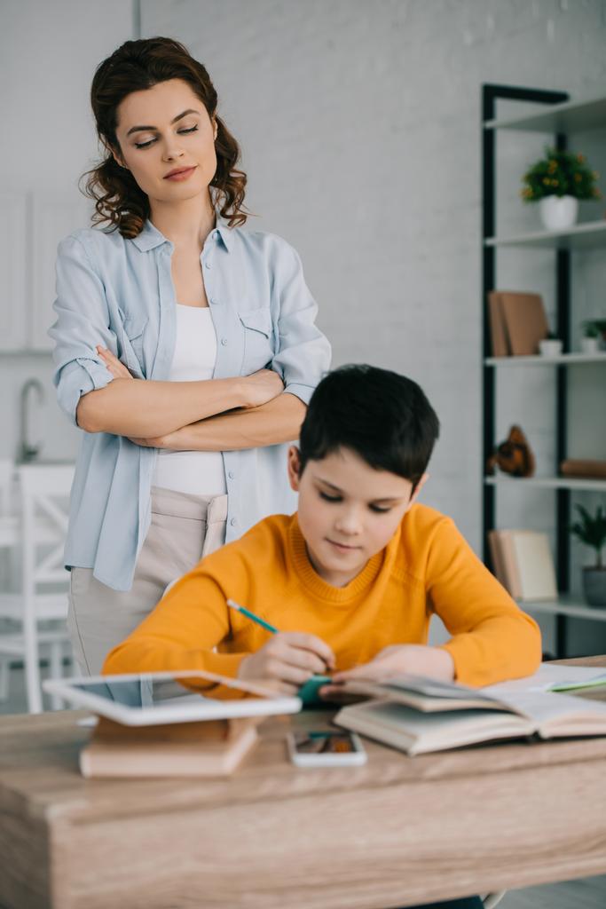 foyer sélectif de mère attentive debout avec le bras croisé derrière le fils assis au bureau et faire des devoirs
 - Photo, image