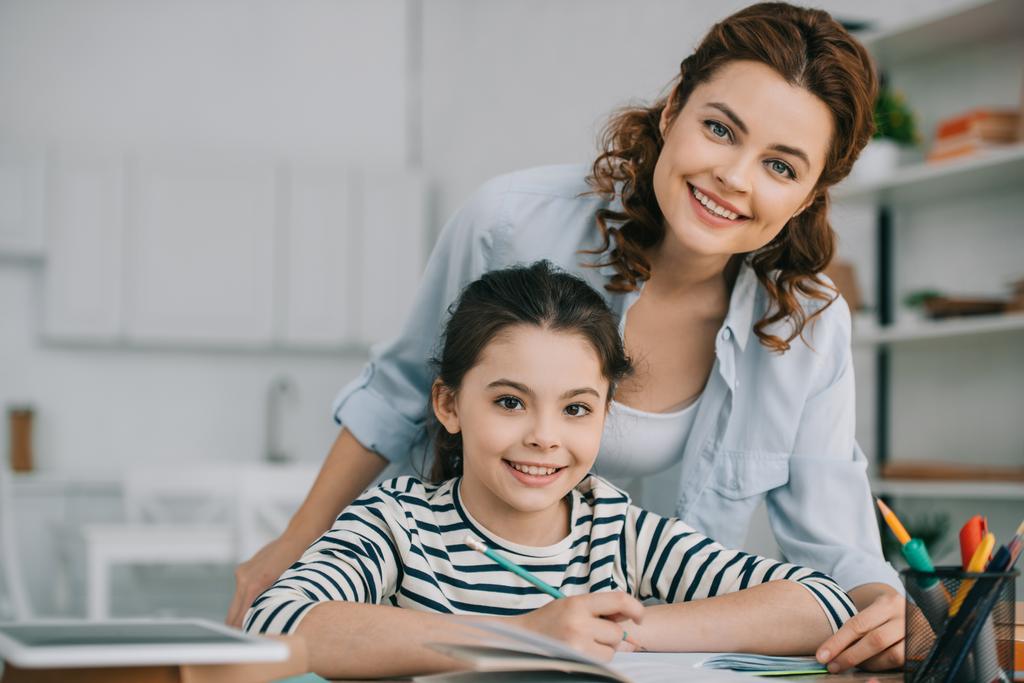happy mother with adorable daughter smiling at camera while doing schoolwork together at home  - Photo, Image