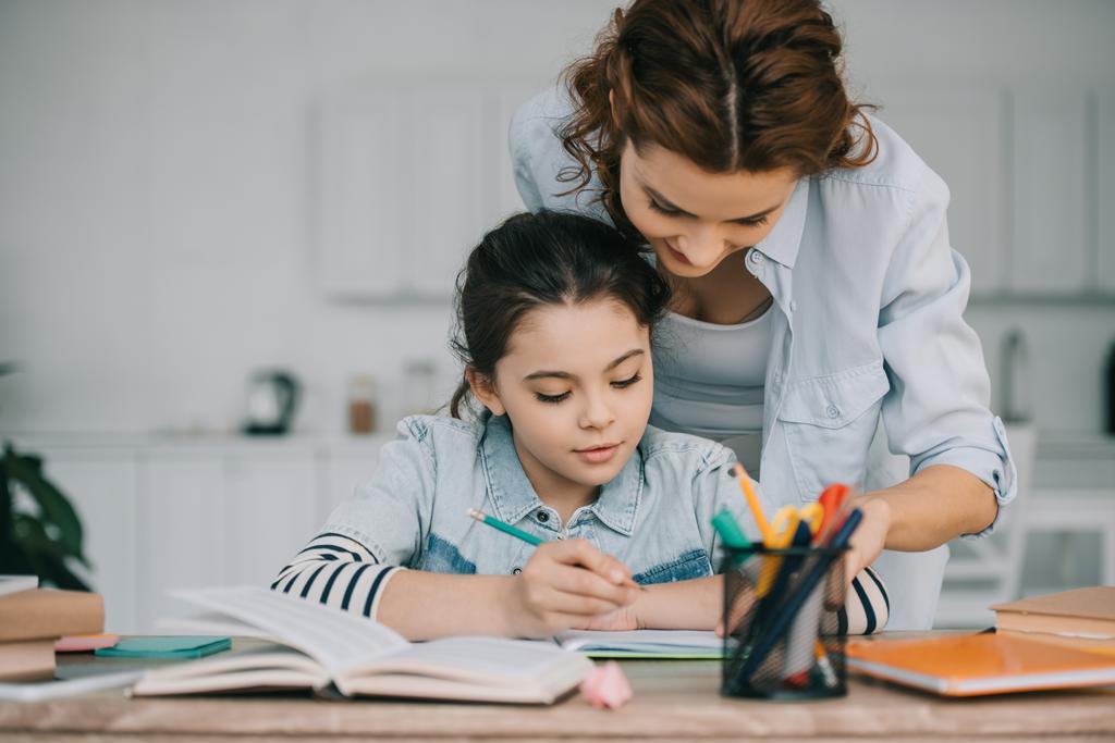 очаровательный ребенок, пишущий в копировальной книге во время школьной работы рядом с матерью
 - Фото, изображение