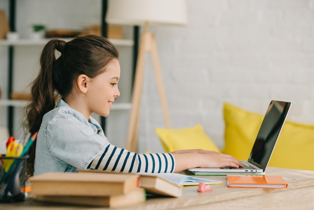 πλευρική όψη του αξιολάτρευτο παιδί χρησιμοποιώντας φορητό υπολογιστή, ενώ κάθονται στο γραφείο με τα βιβλία και να κάνει την εργασία - Φωτογραφία, εικόνα