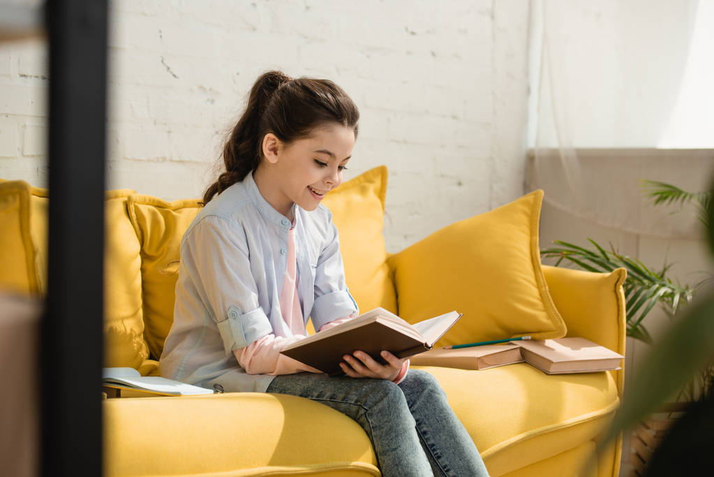 Urocza uśmiechnięta dziecko siedzi na żółtej kanapie i czytając książkę  - Zdjęcie, obraz