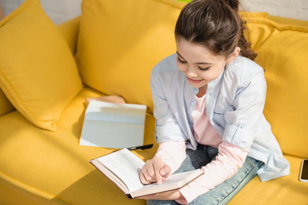 lindo niño sonriente libro de lectura mientras está sentado en el sofá amarillo en casa
 - Foto, imagen