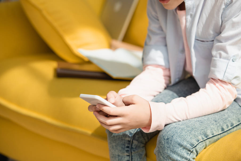 abgeschnittene Ansicht eines Kindes mit Smartphone, während es zu Hause auf dem Sofa sitzt - Foto, Bild