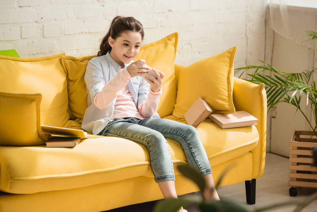 entzückend lächelndes Kind sitzt auf Sofa neben Büchern und benutzt Smartphone - Foto, Bild
