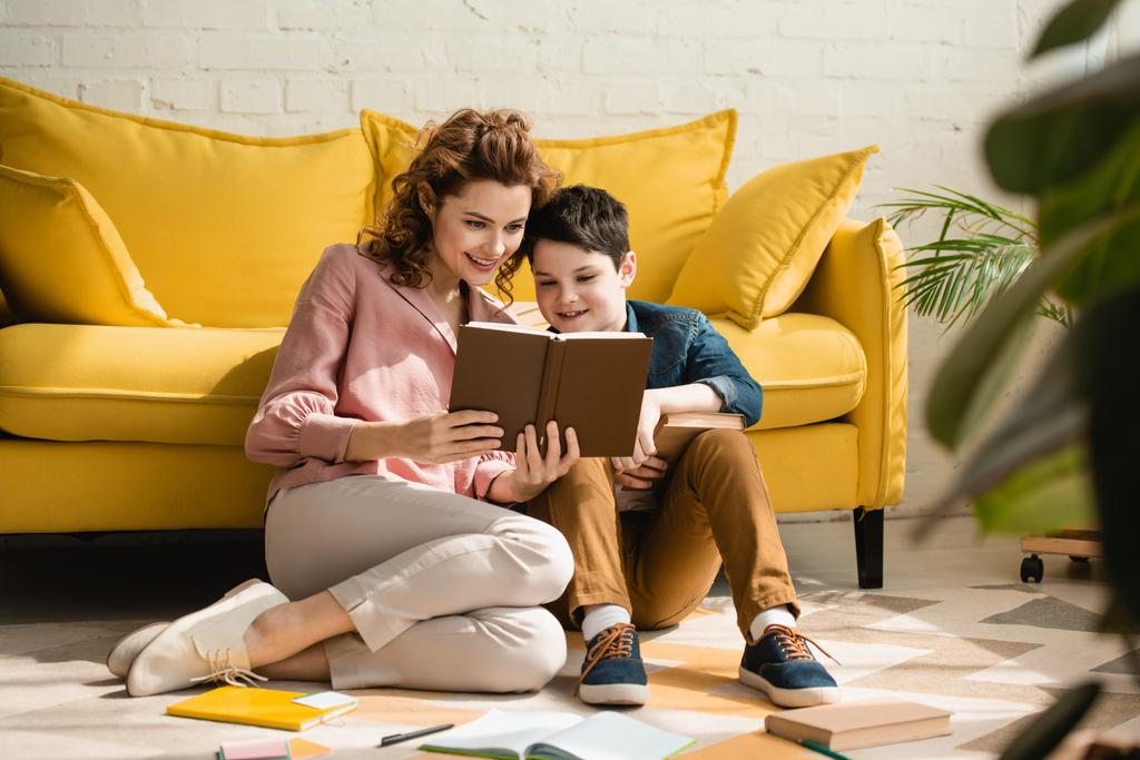 uśmiechnięta matka i syn siedzi na podłodze w pobliżu żółtej kanapie i czytania książki razem - Zdjęcie, obraz