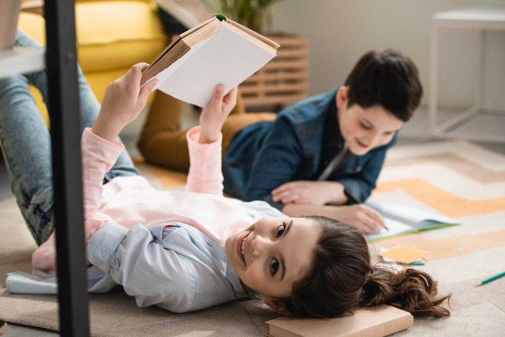selective focus of cheerful child lying on floor with book near brother writing in notebook - Photo, Image