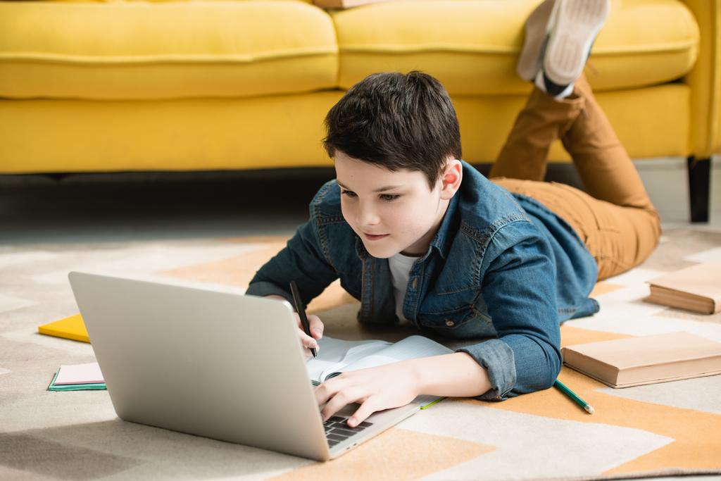uważny chłopiec leżący na podłodze w pobliżu książek i przy użyciu laptopa robiąc pracę domową - Zdjęcie, obraz