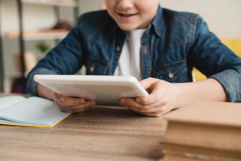 Частичное представление улыбающегося мальчика с помощью цифрового планшета, сидящего за столом с книгами дома - Фото, изображение
