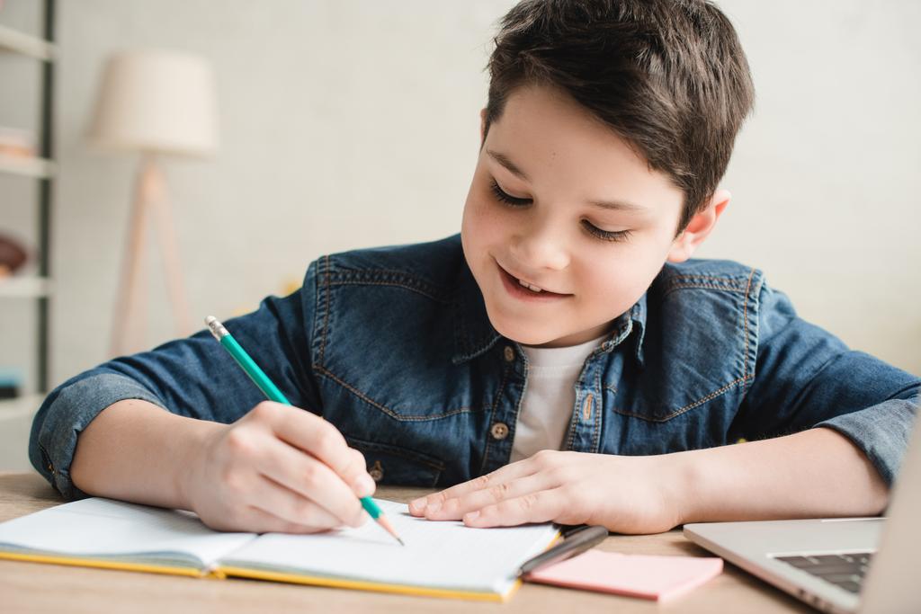 Veselý chlapec, který píše v poznámkovém bloku, sedí u stolu a dělá úkoly - Fotografie, Obrázek