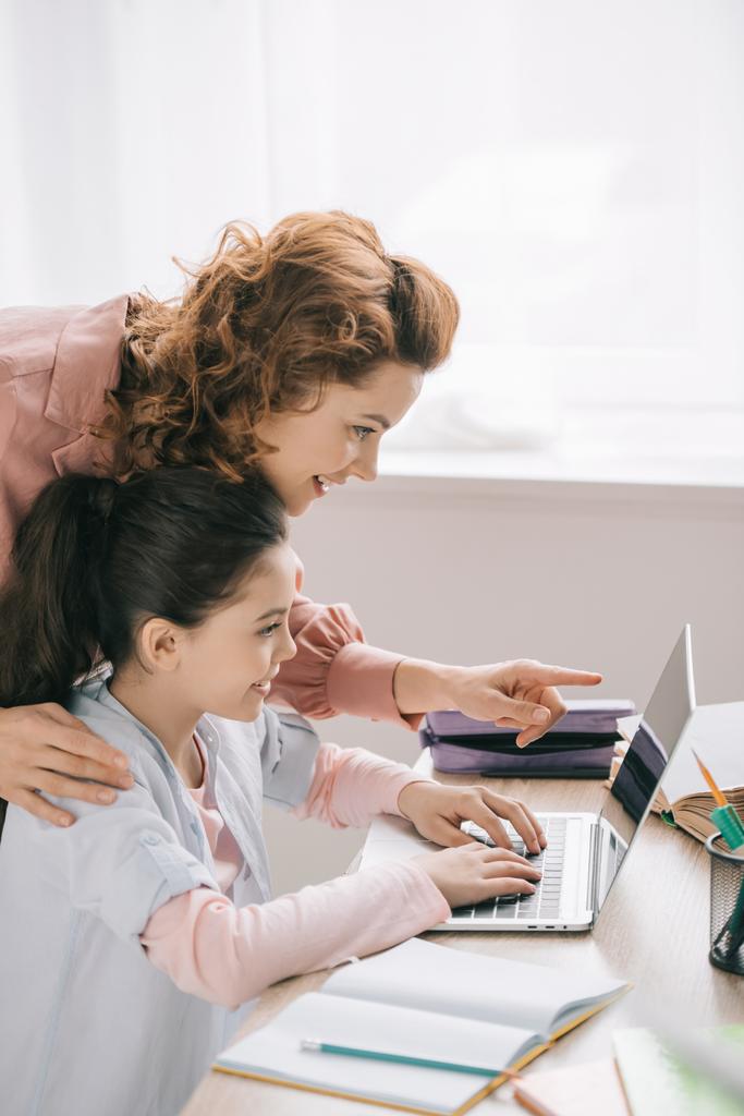smiling mother pointing with finger at laptop screen near adorable daughter doing homework - Photo, Image