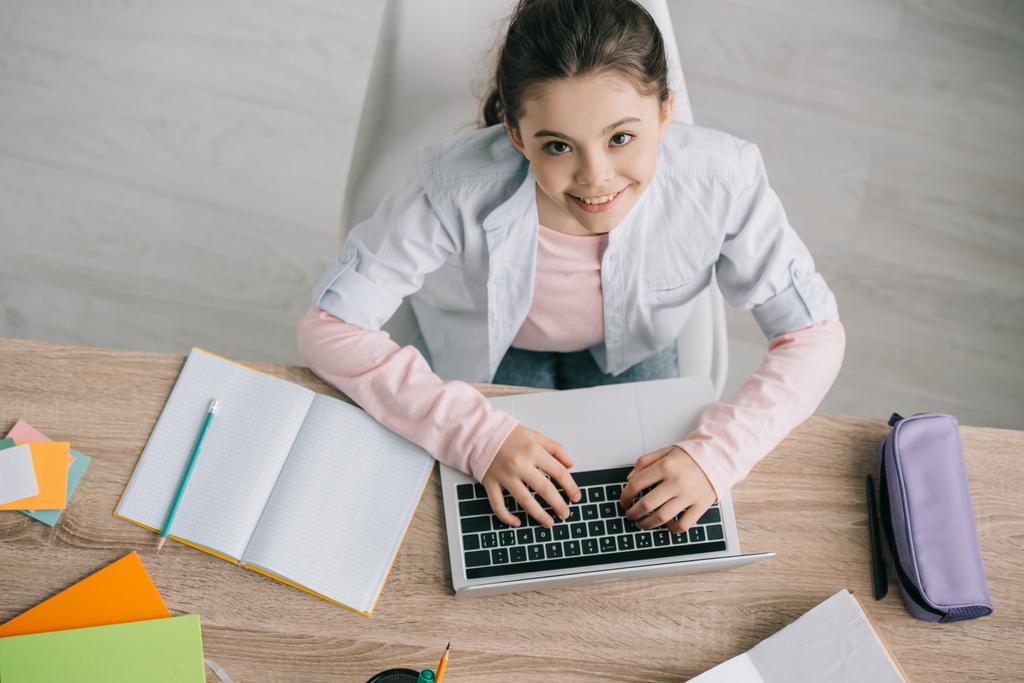 bovenaanzicht van schattig kind met behulp van laptop terwijl zittend op houten bureau en het doen van school werk thuis  - Foto, afbeelding