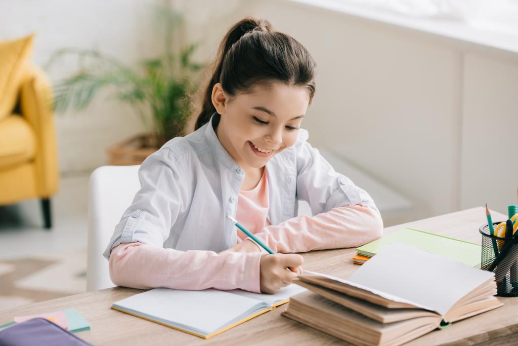 αξιολάτρευτο χαρούμενο παιδί γράφοντας στο σημειωματάριο, ενώ κάθονται στο γραφείο κοντά στο βιβλίο και να κάνει σχολικές εργασίες στο σπίτι - Φωτογραφία, εικόνα