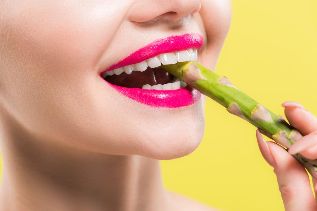 vista cortada de mulher alegre segurando espargos verdes e saborosos na boca isolada no amarelo
  - Foto, Imagem