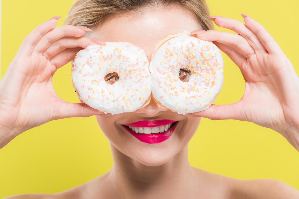 Veselá žena pokrývající oči s chutnými koblihy izolovanými na žluté  - Fotografie, Obrázek