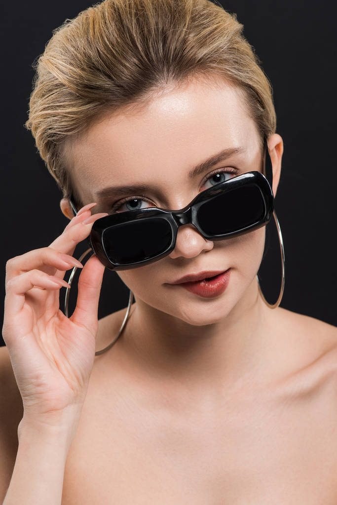 attraktive junge Frau beißt Lippen und berührt Sonnenbrille isoliert auf schwarz  - Foto, Bild