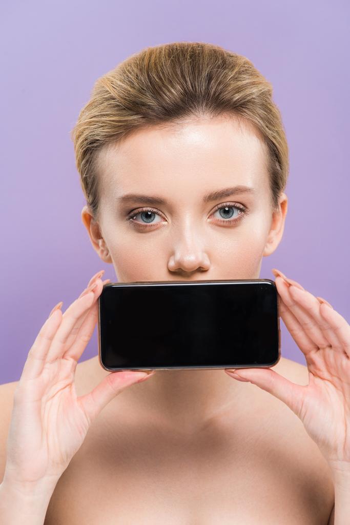 attraktive nackte Frau bedeckt Mund mit Smartphone mit leerem Bildschirm isoliert auf lila   - Foto, Bild