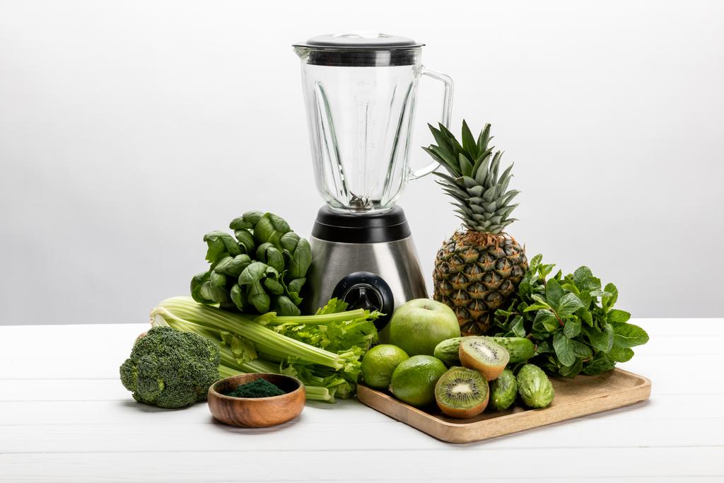 Mixer in der Nähe von grünem frischem Gemüse und leckeren Früchten auf weißem  - Foto, Bild