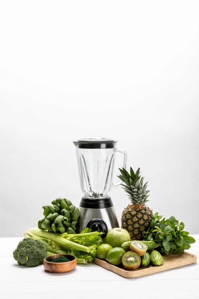 μπλέντερ κοντά σε φρέσκα, πράσινα λαχανικά και νόστιμα φρούτα σε λευκό  - Φωτογραφία, εικόνα