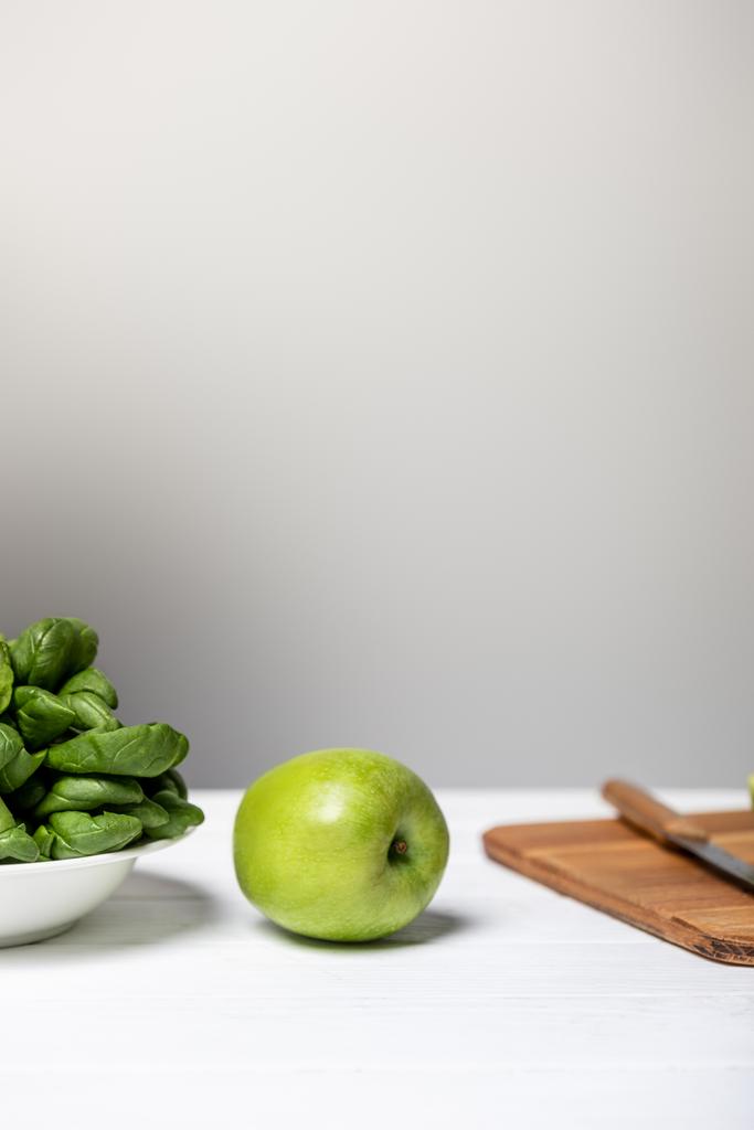 Teller mit Spinatblättern neben grünem Apfel und Schneidebrett auf grau  - Foto, Bild