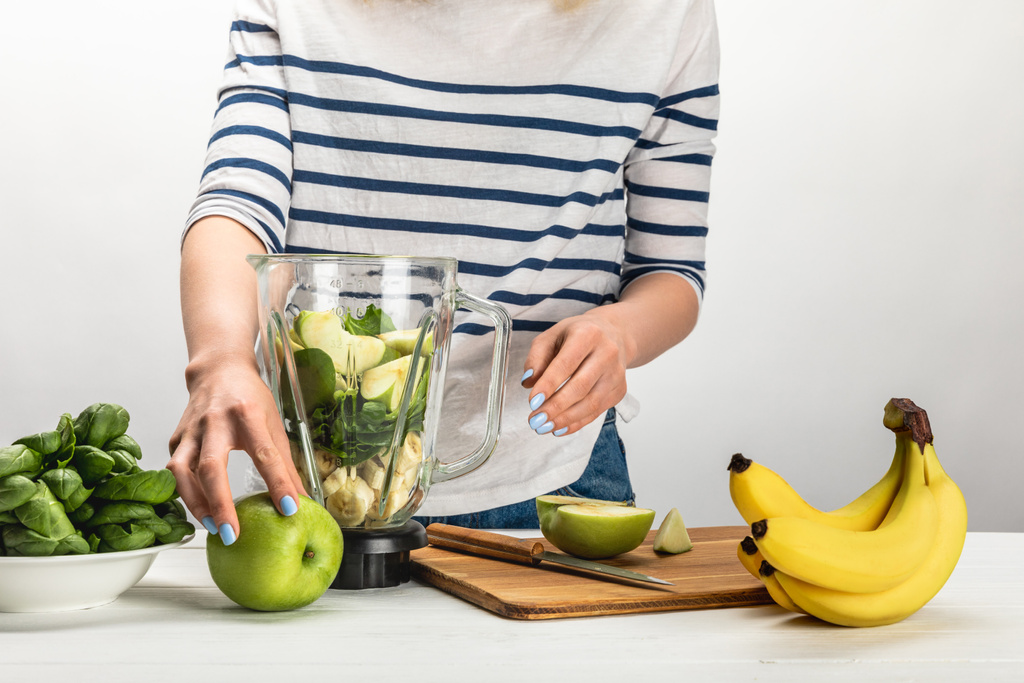 vista recortada de la mujer tomando manzana orgánica cerca de la licuadora con ingredientes en blanco
  - Foto, Imagen
