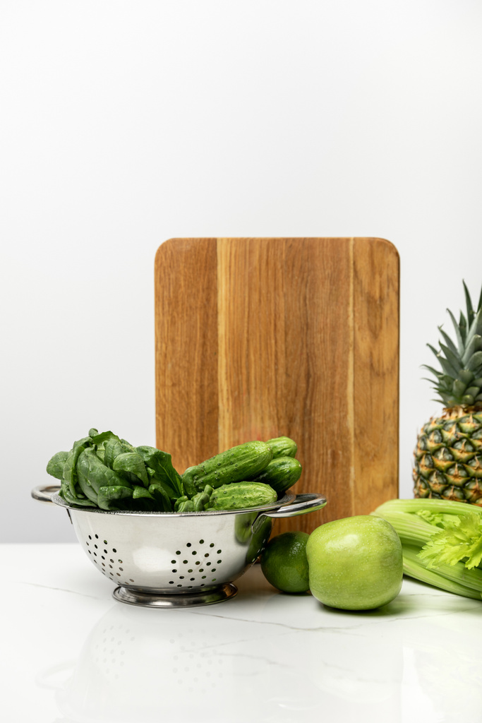 ώριμα φρούτα κοντά σε φρέσκα πράσινα λαχανικά και ξύλινη σανίδα κοπής σε λευκό  - Φωτογραφία, εικόνα