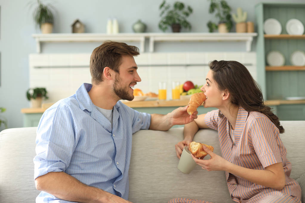 glückliches junges Paar im Schlafanzug in der Küche frühstückt, isst einander ein Croissant. - Foto, Bild
